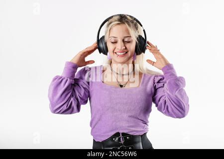 Giovane ragazza bionda sorridente felice indossando lilla coste blusa isolato su sfondo bianco chiuso gli occhi, ascolta la musica preferita, tocca le cuffie Foto Stock