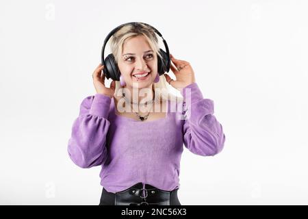 Il ritratto della giovane donna isolato su sfondo bianco tocca le cuffie e ascolta la canzone preferita. Godendolo molto, canta e balla lungo Foto Stock