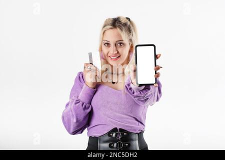Felice donna bionda in piedi isolato su sfondo bianco mostra la fotocamera bianco schermo mockup bianco del telefono e tiene una carta di credito in othe Foto Stock