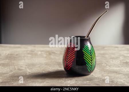 tazza di yerba mate in ceramica piuttosto lucida, con foglie rosse e verdi e paglia di metallo bombarbilla su un tavolo da banco in cemento Foto Stock