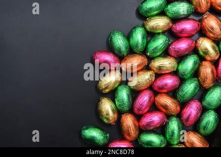 Uova pasquali multicolore avvolte in foglio di colore rosa, verde, arancione e giallo in un palo o in un gruppo, su sfondo grigio nero. Foto Stock