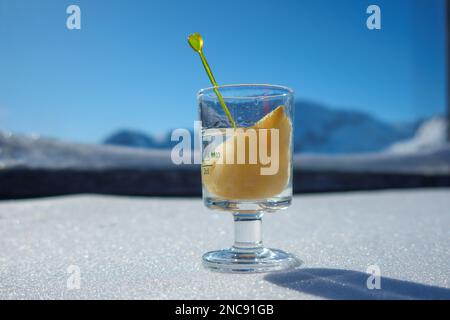 Un bicchiere pieno di schnapps e un pezzo di pera si trova nella neve di fronte alle montagne innevate delle Alpi Foto Stock