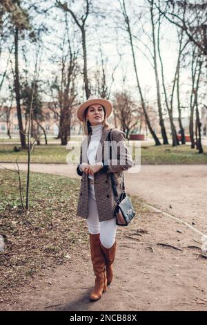 Ritratto a corpo pieno di bella donna che indossa blazer, cappello e stivali eleganti in primavera parco. Moda femminile. Abiti e accessori moderni Foto Stock