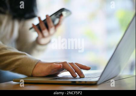 Vista laterale di una donna asiatica freelance che lavora in remoto al bar, utilizzando il computer portatile e il suo smartphone. immagine ritagliata Foto Stock