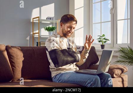 Giovane uomo sorridente con braccio rotto che effettua una videochiamata con un computer portatile Foto Stock