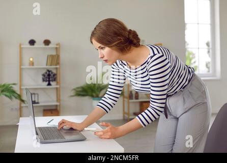 Una giovane e seria donna d'affari freelancer che usa il telefono cellulare e il computer portatile nel suo lavoro in ufficio a casa. Foto Stock