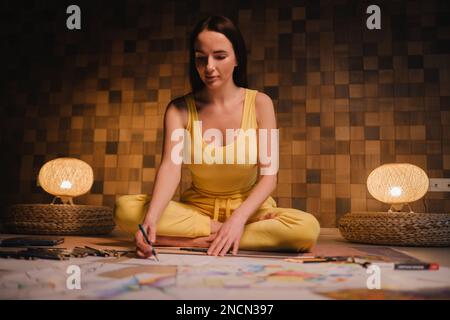 Una ragazza in abiti gialli seduta nella posizione del loto disegna su carta con illuminazione serale a casa Foto Stock