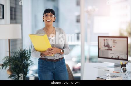 Passando attraverso la mia giornata con un sorriso. Ritratto ritagliato di una giovane donna d'affari attraente in piedi da sola e tenendo i file nel suo ufficio. Foto Stock