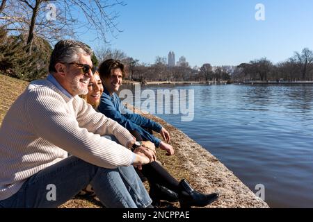 Vista laterale corpo completo di positivo uomo maturo grigio con capelli in occhiali da sole seduti con moglie e figlio vicino al fiume, mentre si passa il fine settimana insieme Foto Stock