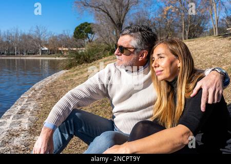 Uomo dai capelli grigi bearded in maglione beige casual e occhiali da sole che abbracciano la moglie bionda mentre si siede sulla riva del fiume contro gli alberi frondosi nel giorno di sole Foto Stock