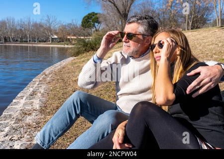 Uomo maturo in abiti casual regolazione occhiali da sole mentre abbraccia moglie indossando vestito nero riposando sulla riva del fiume guardando lontano Foto Stock