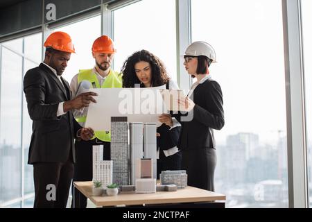 Architetti, ingegneri e designer qualificati che si riuniscono in ufficio di fronte al modello cittadino 3D. Colleghi multiculturali in abiti e caschi in piedi vicino al tavolo e guardando il BluePrint. Foto Stock