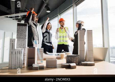 Collaboratori di successo costruttori di team architetti alzando le mani in pugni e urlando con felicità, mentre in piedi tavolo con BluePrint, gadget e la progettazione di edifici residenziale progetto maquette Foto Stock