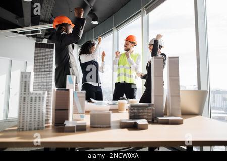 Collaboratori di successo costruttori di team architetti alzando le mani in pugni e urlando con felicità, mentre in piedi tavolo con BluePrint, gadget e la progettazione di edifici residenziale progetto maquette Foto Stock
