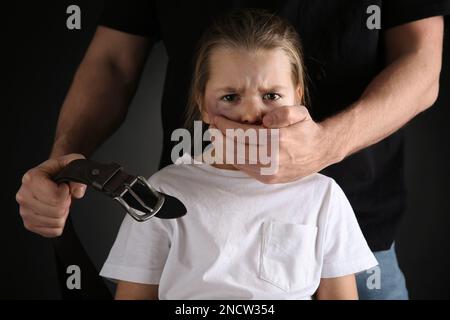 Uomo con cintura che copre la bocca della bambina spaventato su sfondo nero. Violenza domestica Foto Stock