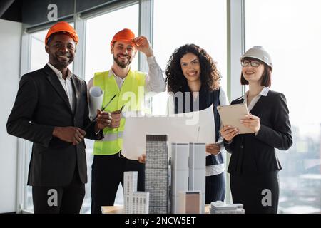 Gruppo di quattro colleghi che si trovano vicino alla costruzione di un progetto prototipo complesso durante una sessione di brainstorming di successo presso l'ufficio dell'ufficio dell'architettura. Squadra di costruzione in piedi con BluePrint e gadget Foto Stock