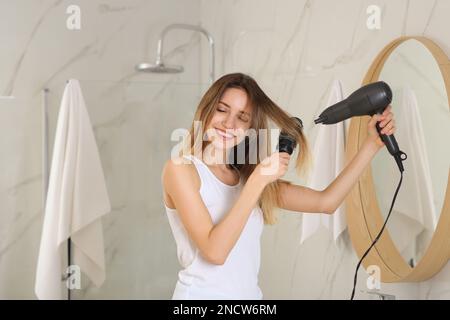 Bella giovane donna che usa l'asciugacapelli vicino allo specchio a casa Foto Stock