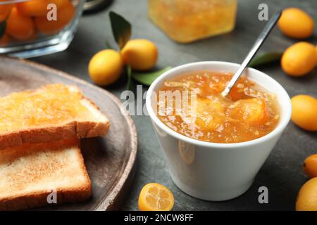 Deliziosa marmellata di kumquat in ciotola, toast e frutta fresca sul tavolo grigio, primo piano Foto Stock