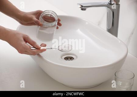 Donna che usa bicarbonato di sodio per disintasare lo scarico del lavandino in bagno, primo piano Foto Stock