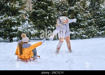 Fiaba invernale, madre felice che tira la slitta con la bambina sulla strada innevata del parco in neve fresca profonda. Godetevi una giornata bianca d'inverno. Passare del tempo insieme Foto Stock