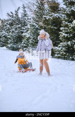 Fiaba invernale, madre felice che tira la slitta con la bambina sulla strada innevata del parco in neve fresca profonda. Godetevi una giornata bianca d'inverno. Passare del tempo insieme Foto Stock