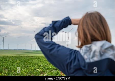 Donna con capelli marroni e giacca blu sta guardando un parco eolico da lontano, parco eolico con turbine eoliche in campo agricolo, vista posteriore Foto Stock