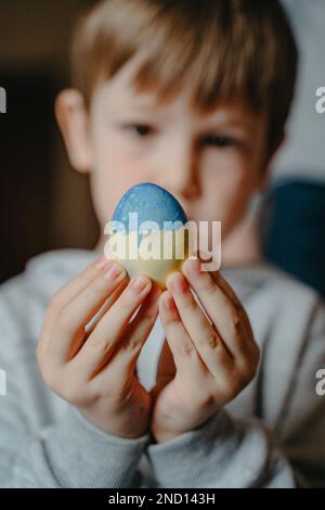 Ragazzino ucraino con un uovo di pasqua dipinto di giallo blu nelle mani, fuoco selettivo sulla parte anteriore Foto Stock