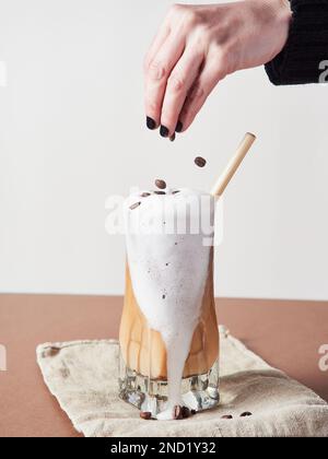 Persona irriconoscibile che aggiunge chicchi di caffè in un bicchiere di delizioso caffè con panna montata e paglia sul tovagliolo in studio Foto Stock