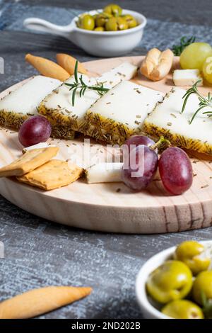 Formaggio appetitoso servito su tavola di legno con uva matura e cracker decorati da rametti di rosmarino vicino alle olive in ciotole sul tavolo Foto Stock