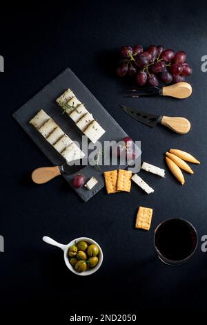 Vista dall'alto di un delizioso formaggio assortito sistemato su un pannello nero con uva matura e cracker collocati vicino a coltelli su una superficie scura Foto Stock