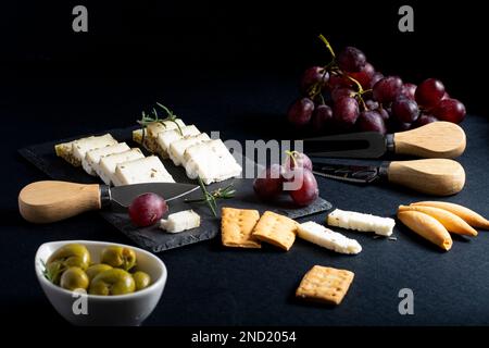 Vista dall'alto di un delizioso formaggio assortito sistemato su un pannello nero con uva matura e cracker collocati vicino a coltelli su una superficie scura Foto Stock