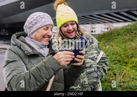 Nonna matura positiva in outerwear e cappello sorridendo e mostrando smartphone a nonna mentre camminano insieme sulla strada Foto Stock