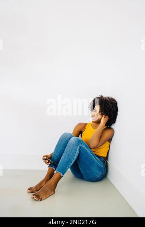Corpo pieno di giovane afro-americana a piedi nudi pensiva in abbigliamento casual con capelli ricci seduti sul pavimento in angolo e guardando di nuovo la macchina fotografica Foto Stock