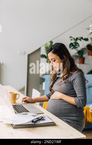 Vista laterale di giovane donna incinta concentrata freelancer con lunghi capelli ondulati seduti a tavolo mentre si lavora a progetto remoto utilizzando il computer a casa Foto Stock