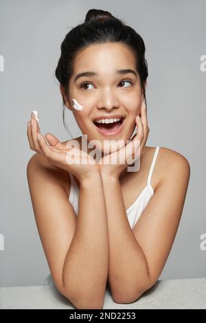 Giovane femmina positiva in cima bianca guardando via mentre si applica crema idratante sul viso Foto Stock