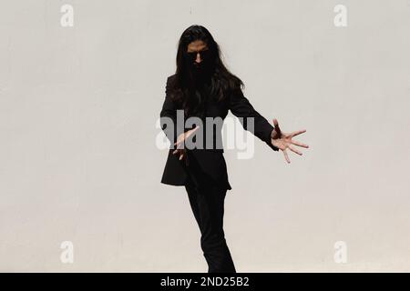Uomo ispanico con capelli lunghi in tuta nera con braccio di sollevamento abiti scuri e flamenco danzante contro parete bianca Foto Stock