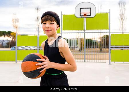 Giovane bambino sportivo in abbigliamento sportivo e cappello dribbling palla mentre si pratica il basket in campo contro cerchio durante l'allenamento Foto Stock