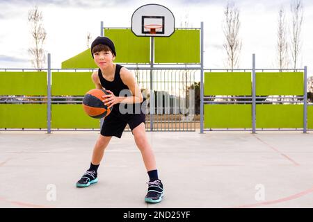 Corpo pieno di giovane bambino sportivo in abbigliamento sportivo e cappello dribbling palla mentre si pratica il basket sul campo contro cerchio durante l'allenamento Foto Stock