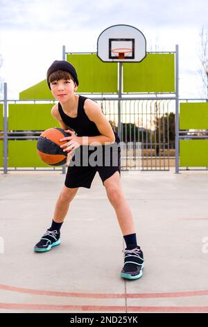 Corpo pieno di giovane bambino sportivo in abbigliamento sportivo e cappello dribbling palla mentre si pratica il basket sul campo contro cerchio durante l'allenamento Foto Stock