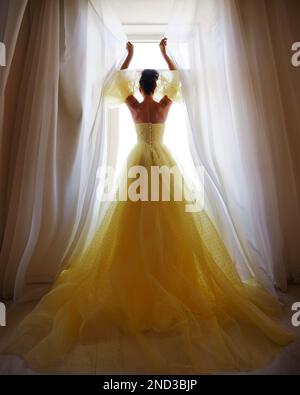 La silhouette di una donna in un abito dorato di lusso sullo sfondo di una finestra tiene una tenda con le mani. Elegante donna in seta lunga gialla Foto Stock