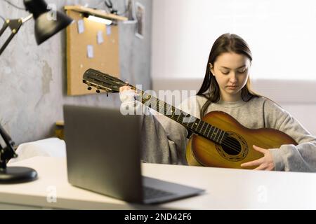 Formazione online, corsi online. Ragazza asiatica focalizzata a suonare la chitarra e a guardare il corso online sul laptop mentre si pratica a casa. Foto Stock