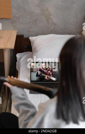 Ragazza teen focalizzata a suonare la chitarra acustica e a guardare il corso online sul laptop mentre si pratica a casa. Formazione online, corsi online. Foto Stock