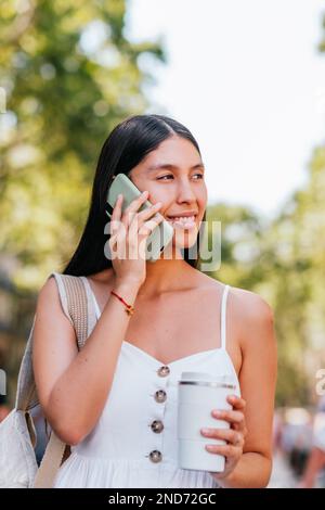 Felice latinoamericana femmina con zero rifiuti takeaway tazza di caffè sorridente e guardando via mentre si parla sul cellulare su sfondo sfocato del parco in estate a Barcellona, Spagna Foto Stock