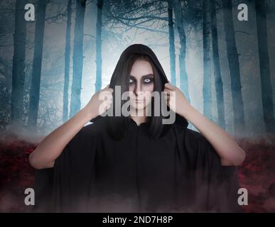 Strega che indossa il mantello nero in una foresta nebbiosa. Carattere fantasy spaventoso Foto Stock