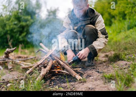 Primo piano di un maschio sopravvissuto sfocato in un impermeabile che inizia il fuoco utilizzando un accendino a gas sulla riva del fiume per cucinare e riscaldare la sera prima del tramonto. Foto Stock