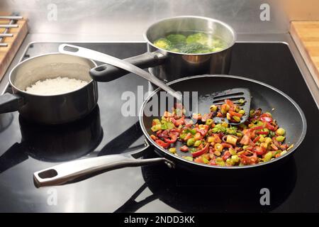 Le verdure tagliate a dadini vengono fritte in padella con spezie, pentole con riso e broccoli dietro di esse sul piano cottura nero, cucina asiatica, copia Foto Stock