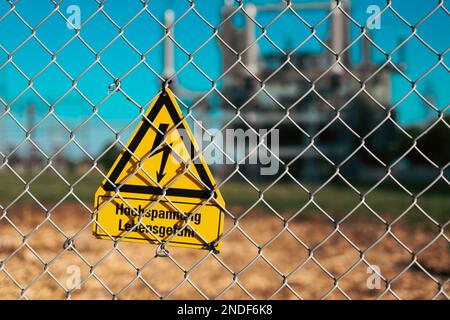 Cartello giallo pericolo alta tensione pericolo di vita su recinzione metallica di fronte a una sottostazione elettrica in Germania Foto Stock