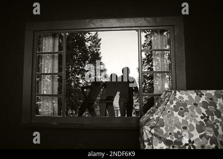 Selfie monocromatico di tre persone riflesse in una finestra di un cottage estivo. Con un barbecue coperto nell'angolo. Luglio 1977 Foto Stock