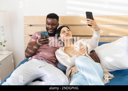 Felice coppia diversa sdraiata a letto e utilizzando gli smartphone. Trascorrere il tempo di qualità a casa, la vita domestica e il concetto di stile di vita. Foto Stock