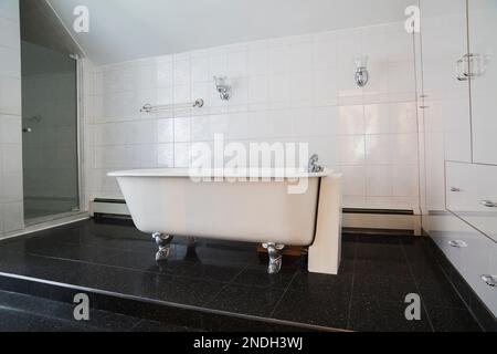 Vasca da bagno con zoccolo in ghisa bianca su piattaforma in piastrelle di granito nero rialzata nel bagno principale con armadietti e cassetti bianchi nella vecchia casa del 1900. Foto Stock